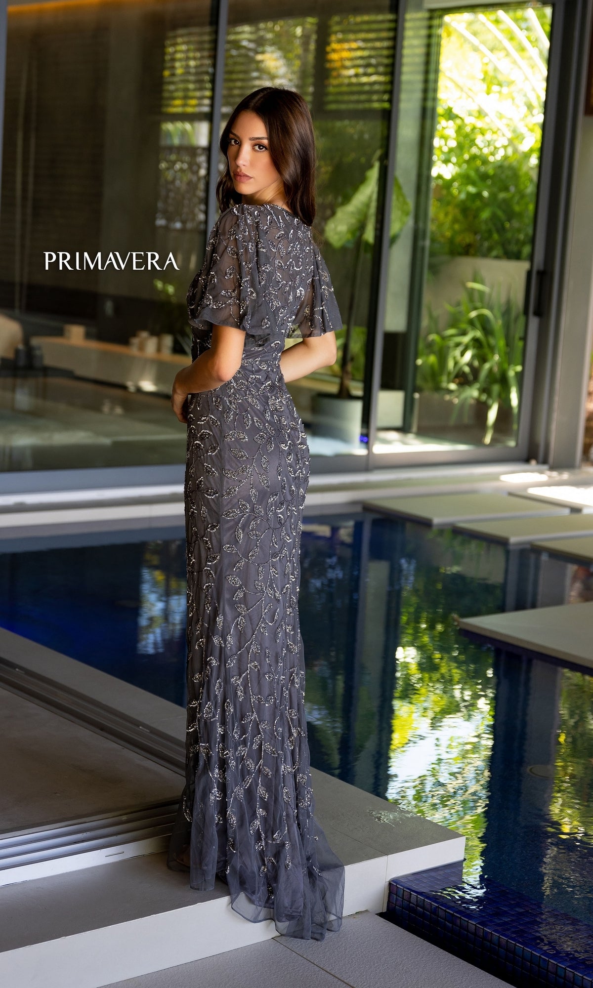A woman in the Long Prom Dress 13111 by Primavera, featuring a high neck and beaded details, stands by an indoor pool looking over her shoulder.